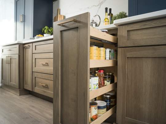 A kitchen with a wooden cabinet that has a pull-out pantry filled with various food items, including bottles and oranges, and a sink on the right.