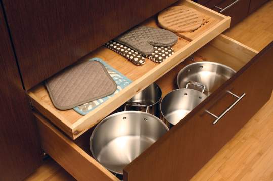 A neatly organized wooden kitchen cabinet containing five pans of various sizes and three oven mitts of different sizes.